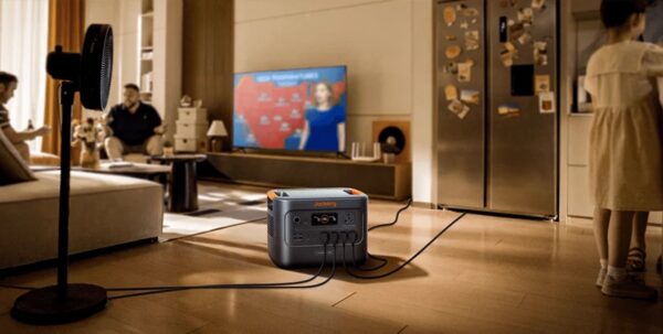A Jackery portable power station on a living room floor powering a fan and TV during a home backup scenario