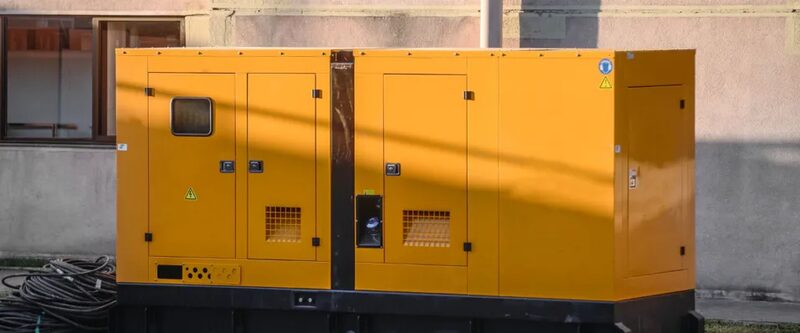 A large yellow industrial standby diesel generator enclosure standing outdoors against a building wall