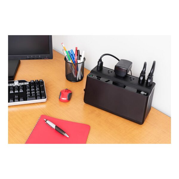 An APC Back-UPS battery backup unit with NEMA outlets sitting on a wooden desk beside a keyboard, monitor and mouse