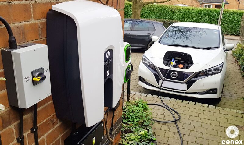 A wall-mounted EV charge point on a brick house with a cable charging a white Nissan Leaf parked on a driveway