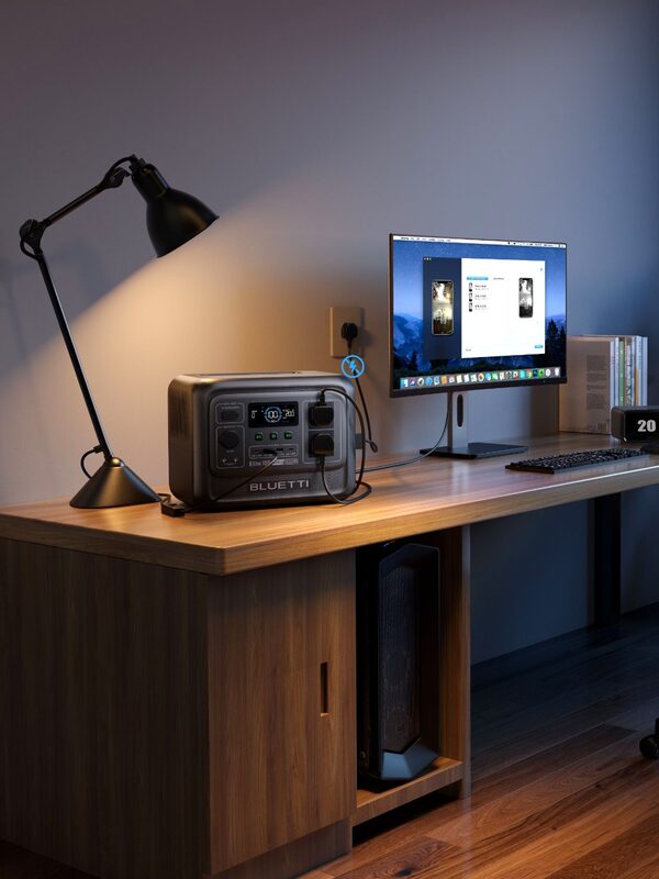 A Bluetti portable power station on a wooden desk powering a desk lamp and computer monitor in a home office