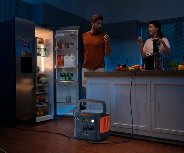 A Jackery portable power station on a kitchen floor powering appliances during a power cut, with a couple at the counter and an open fridge