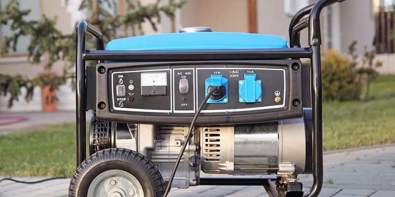 A portable petrol generator with a blue fuel tank, control panel with 230V sockets and wheels, standing outdoors on a paved drive