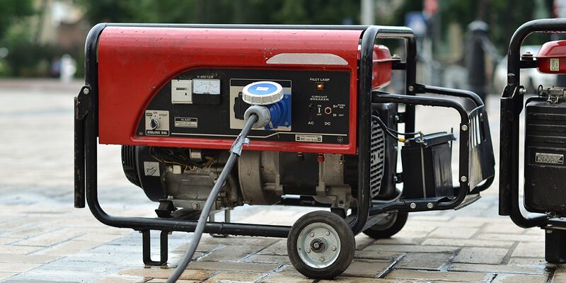 A red portable petrol generator on wheels with a blue plug socket and cable, standing outdoors on paving