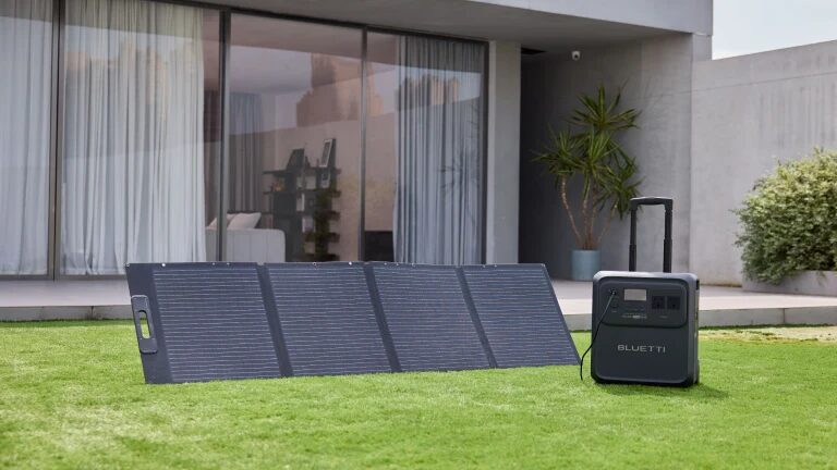 A Bluetti portable power station with a folding solar panel set up on a lawn outside a modern house