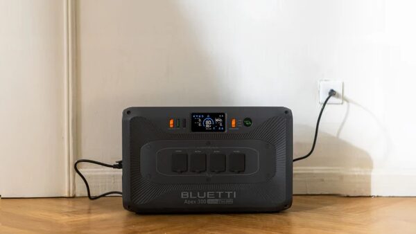 A Bluetti Apex 300 portable power station plugged into a wall socket on a wooden floor