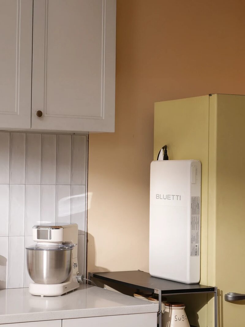 A white BLUETTI portable power station standing on a shelf in a kitchen next to a fridge and a stand mixer