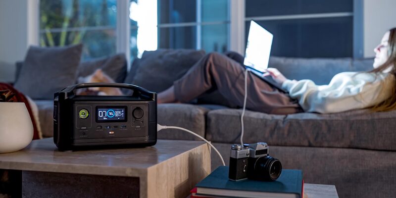 A black EcoFlow portable power station on a coffee table charging a laptop used by a woman on a sofa
