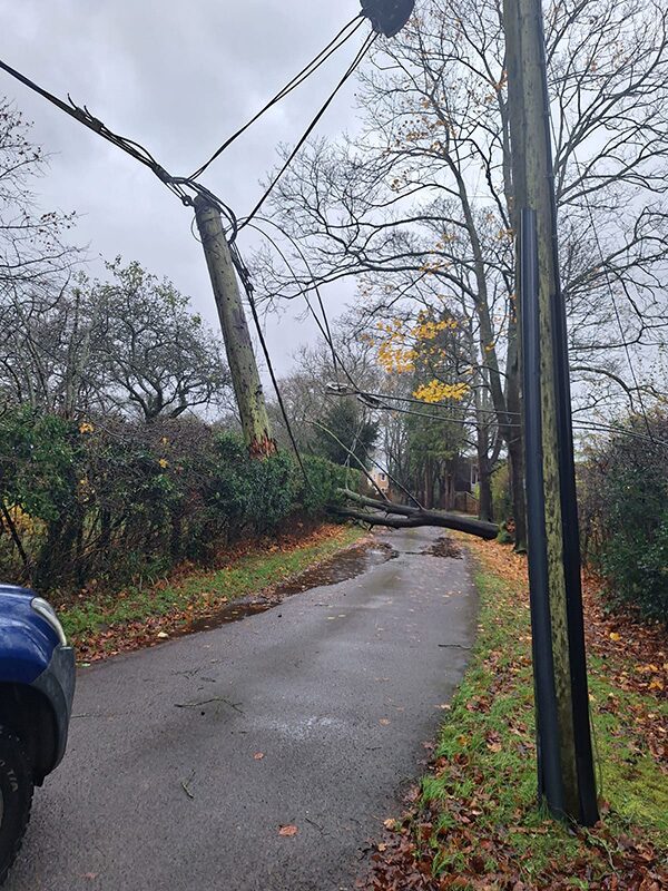 A storm-damaged wooden electricity pole leaning over a wet rural road with fallen power lines and construction equipment in the background