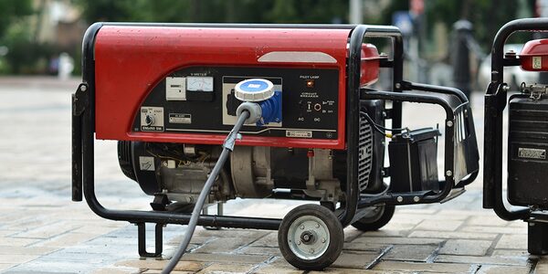 A red portable petrol generator on wheels with a blue plug socket and cable, standing outdoors on paving