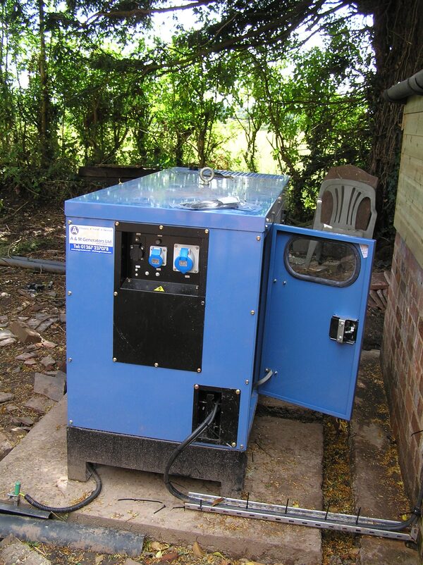 A blue standby generator cabinet installed outdoors on a concrete slab beside a brick building, with its door open showing sockets and controls