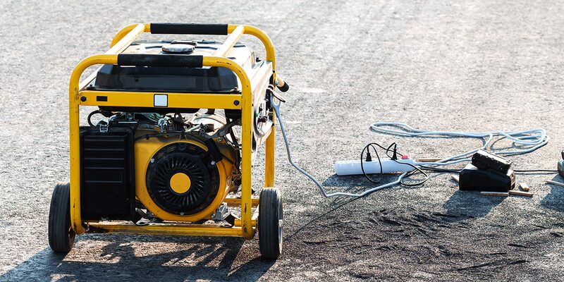 A yellow portable petrol generator with wheels and a pull cord standing on tarmac, with cables and tools lying beside it