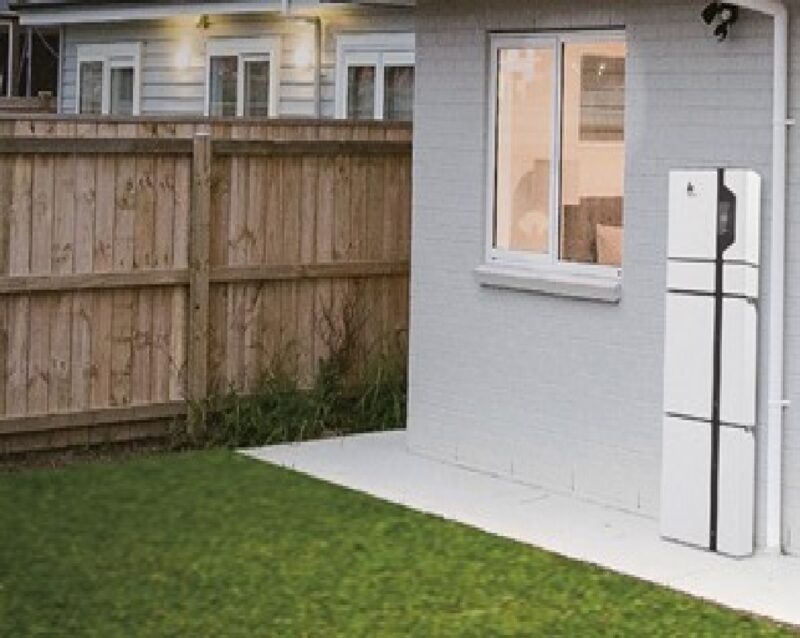 A white home battery storage unit mounted on the exterior wall of a house beside a window, with a lawn and wooden fence in the foreground