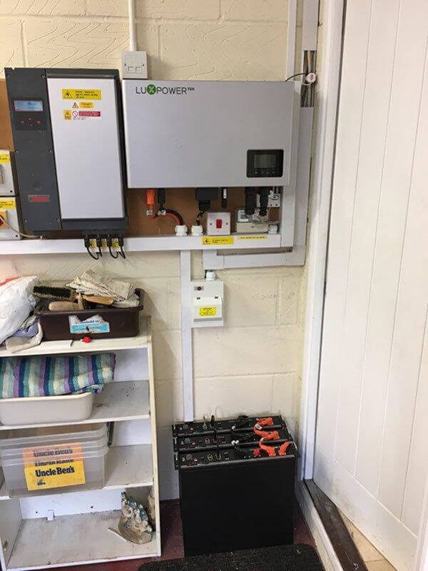 A LuxPowerTek hybrid inverter and battery storage unit installed on a wall next to a consumer unit, with batteries on the floor below