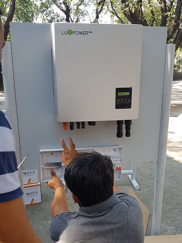 A LuxPowerTek hybrid solar inverter mounted on an outdoor panel while a technician works on the wiring below