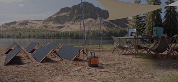 Jackery portable power station charging from fold-out solar panels at a lakeside campsite with tent and camping chairs