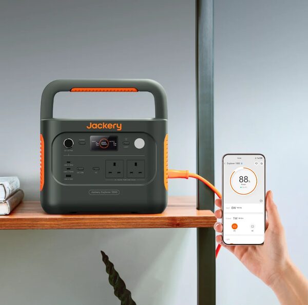 A Jackery Explorer 1000 portable power station on a shelf with a hand holding a phone showing its app