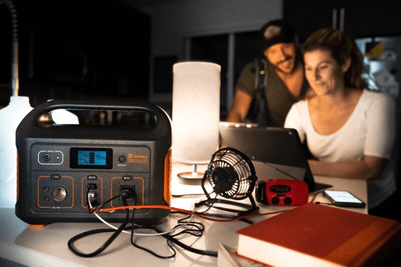 A Jackery portable power station on a table powering a fan and lamp while a couple use a laptop behind it
