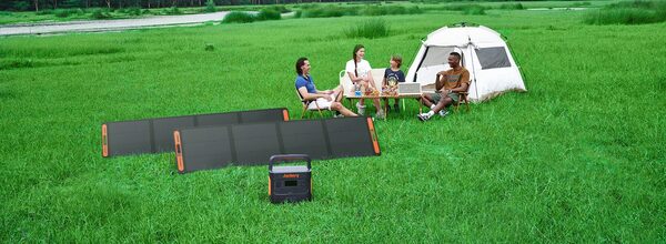 A Jackery portable power station with foldable solar panels on grass beside a family camping by a tent