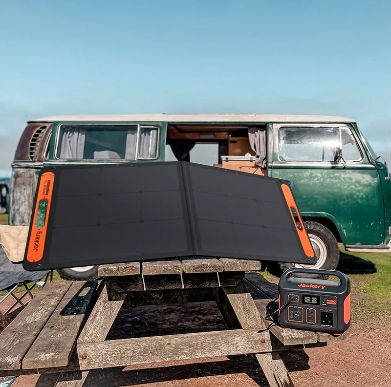 A Jackery portable solar panel and Explorer 500 power station set up on a wooden picnic table in front of a green camper van