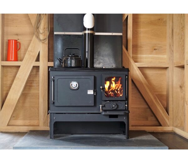 A black wood-burning cook stove with a lit firebox and a kettle on top, installed in a timber-framed room