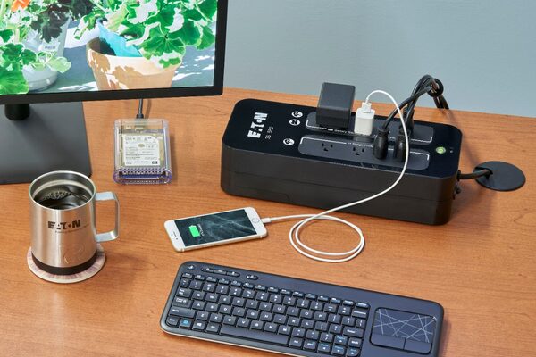 An Eaton 3S UPS battery backup unit on a wooden desk powering a monitor, phone charger and other devices