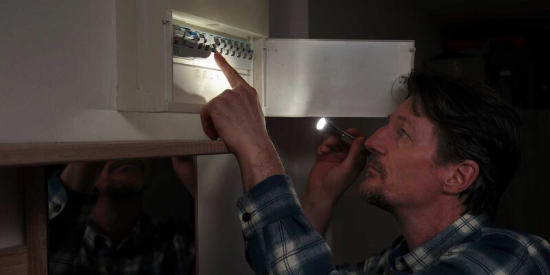 A man holding a torch inspects a wall-mounted consumer unit (fuse box) with its cover open