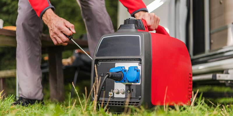 A person pulling the starter cord on a red portable inverter generator set up on grass next to a caravan