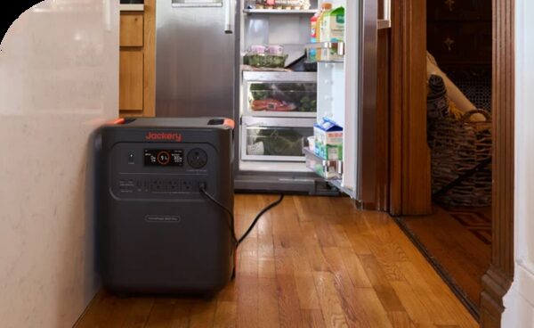 A black Jackery portable power station plugged in on a kitchen floor next to an open fridge