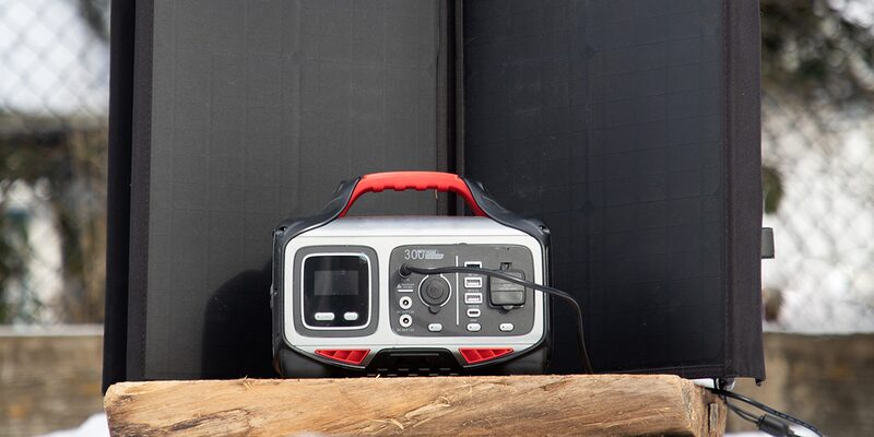 A portable power station with a red handle sitting on a wooden beam in front of a folding solar panel outdoors