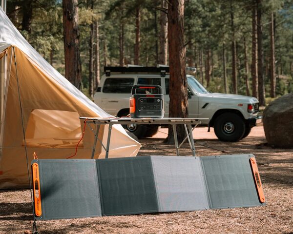 A Jackery portable power station on a camping table connected to a folding portable solar panel at a campsite with a tent and 4x4 vehicle