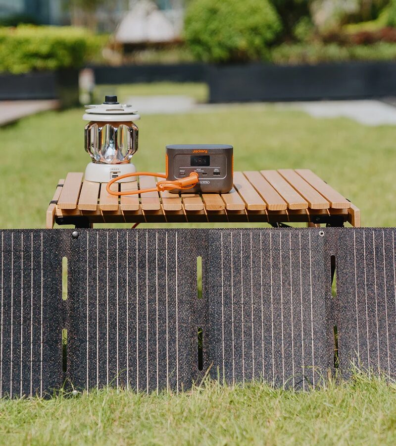A Jackery portable power station and folding solar panel set up on a garden lawn with a camping lantern on a wooden table
