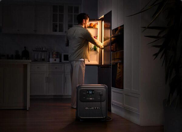 A Bluetti portable power station on a kitchen floor powering a fridge while a man opens the fridge door in a dark kitchen during a power cut