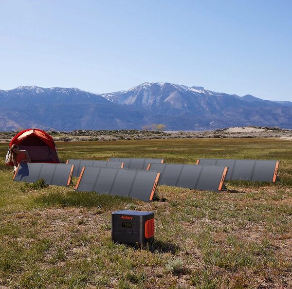A Jackery portable power station charging from several folding solar panels set up on grass at a campsite with a tent and mountains behind