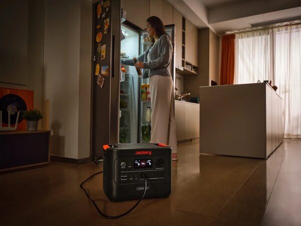 A Jackery HomePower 3000 portable power station on a kitchen floor powering a fridge while a woman opens the fridge door