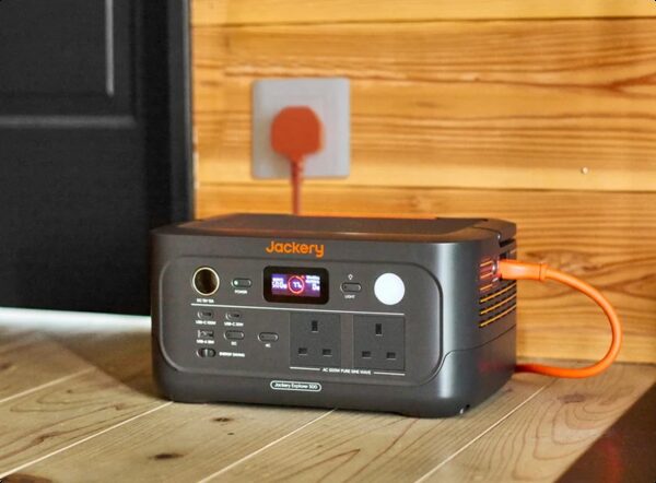 A Jackery Explorer 500 portable power station plugged into a UK wall socket with an orange charging cable