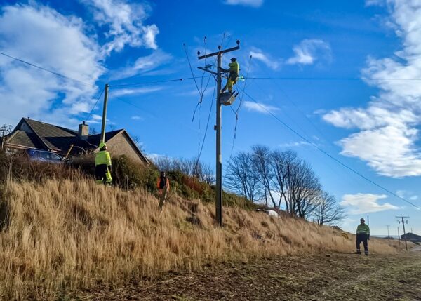 Electricity engineers in high-visibility clothing repairing a damaged wooden power pole in the countryside near a house after a storm
