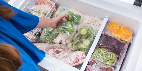 A woman reaching into an open chest freezer full of bags of frozen fruit, vegetables and meat