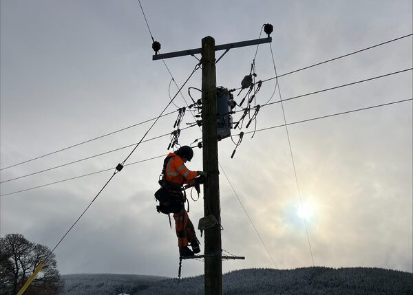 An engineer in orange high-visibility safety gear climbing a wooden electricity distribution pole to repair power lines against a wintry sky