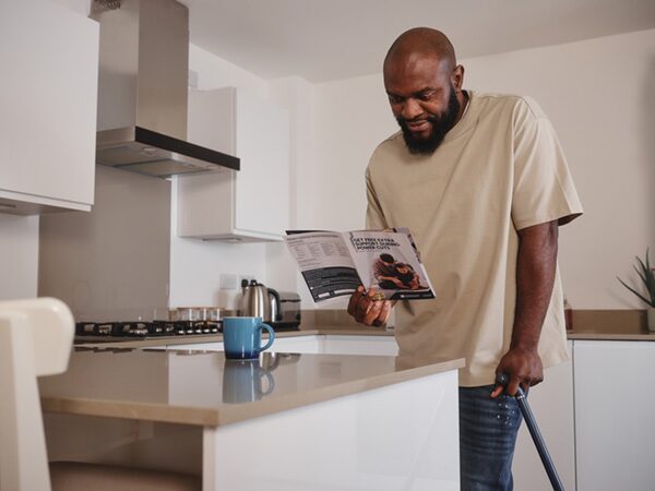A man with a walking stick standing in a kitchen reading a Priority Services Register leaflet