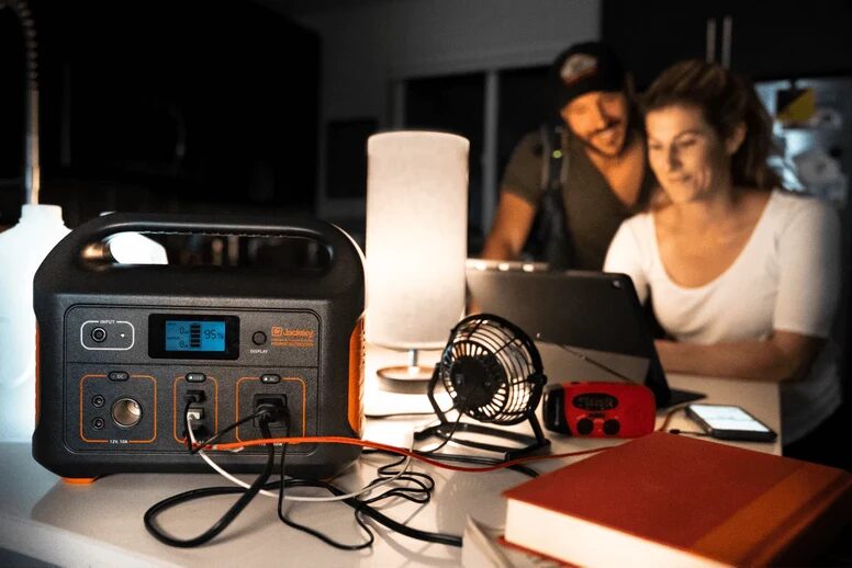 A Jackery portable power station on a table powering a fan and lamp while a couple use a laptop in the background