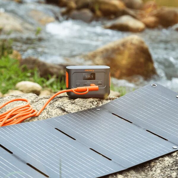 A Jackery portable power station charging from a folding solar panel on rocks beside a stream