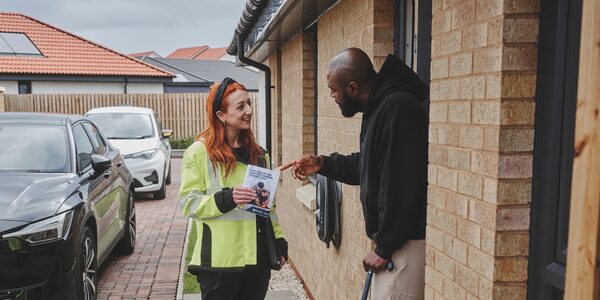 A SSEN worker in a high-visibility jacket handing a Priority Services Register leaflet to a man at his front door on a residential driveway