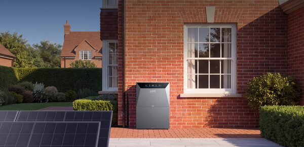An Anker SOLIX Solarbank battery unit installed on the exterior wall of a brick house with solar panels in the foreground
