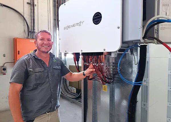 A smiling installer standing next to a wall-mounted Sigenergy home battery inverter unit with exposed wiring in a utility space
