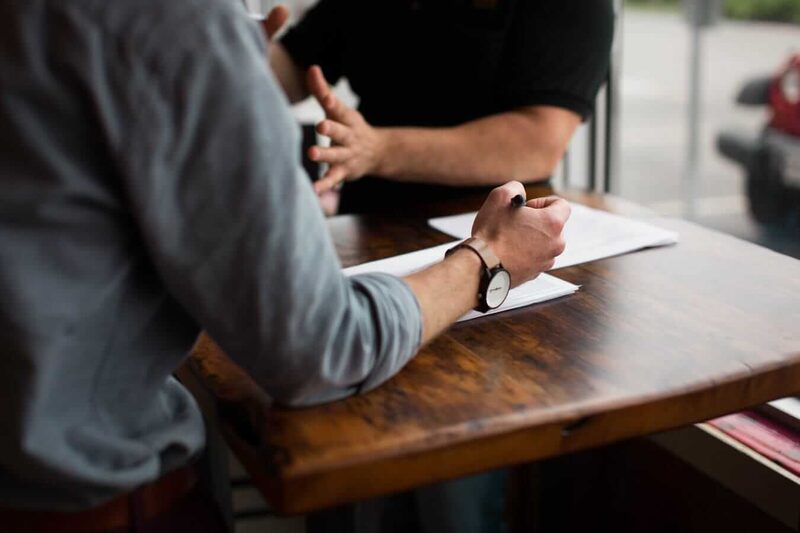 Two people in discussion over paperwork at a wooden table, one writing on documents