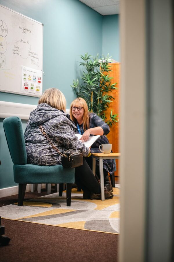 An adviser with a lanyard helping a seated woman with paperwork in a small consultation room with a whiteboard