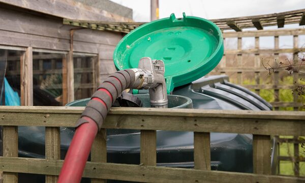 A red delivery hose filling a green domestic heating oil tank behind a wooden fence