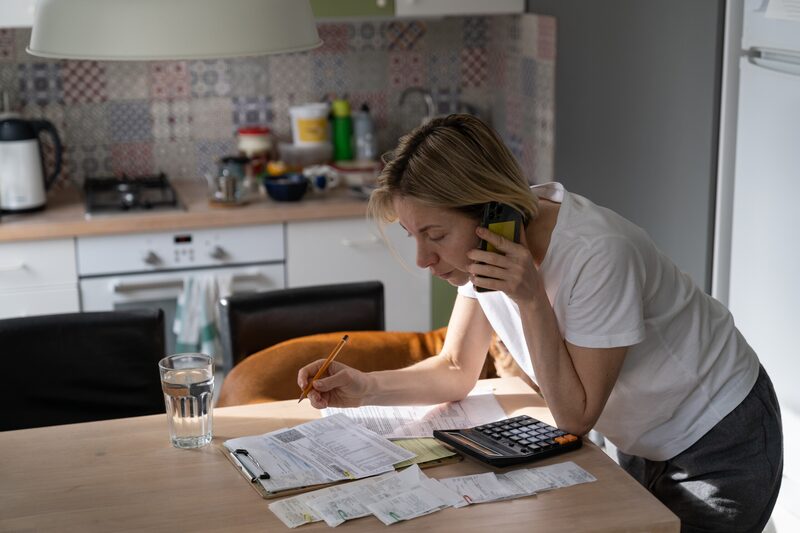 A woman on the phone at a kitchen table working through bills with a calculator and pencil