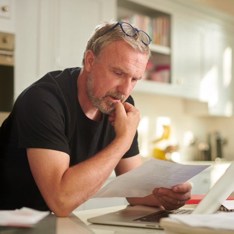 A middle-aged man at a kitchen table reading a paper bill next to an open laptop